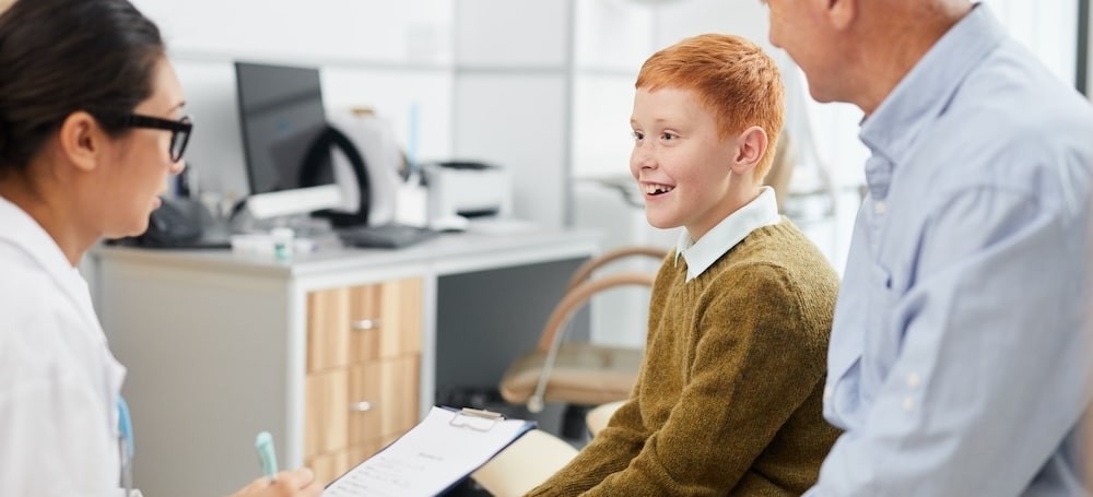 Side view portrait of smiling boy visiting doctor with father and looking at nurse filling patients form in waiting room