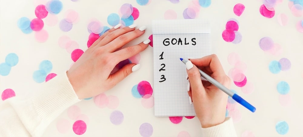 Female hand with pen writing goals in empty notebook on white background with confetti