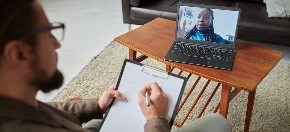 High angle veiw of psychologist making notes on paper while talking to patient online on laptop during psychotherapy session